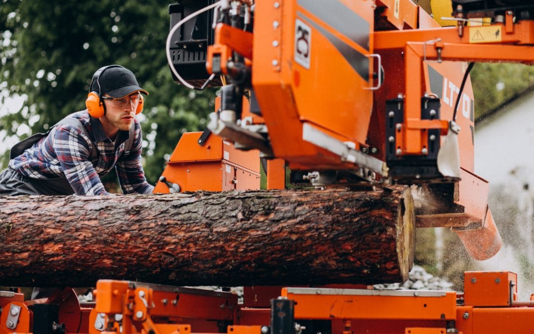Man wearing ear protection operates an orange sawmill cutting a large log outdoors.