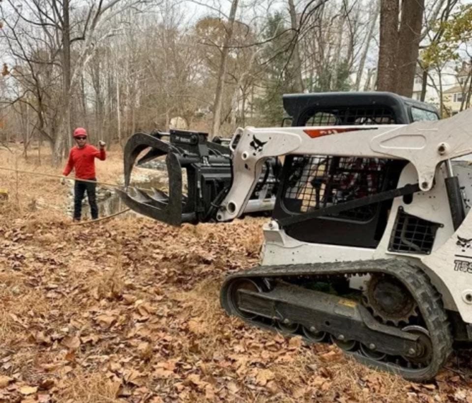 damage-img Person in red helmet stands near a skid steer loader with a grapple attachment in a wooded area.