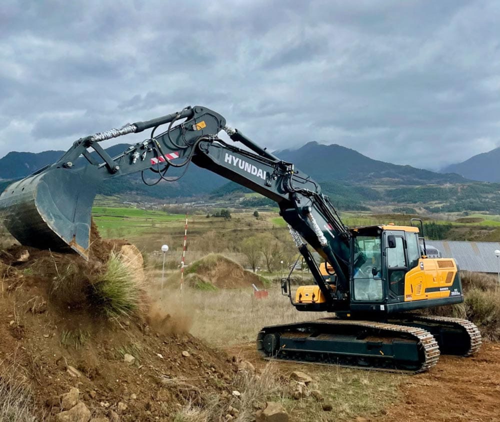 lot-clean-img A Hyundai excavator lifts dirt on a construction site with mountains in the background.